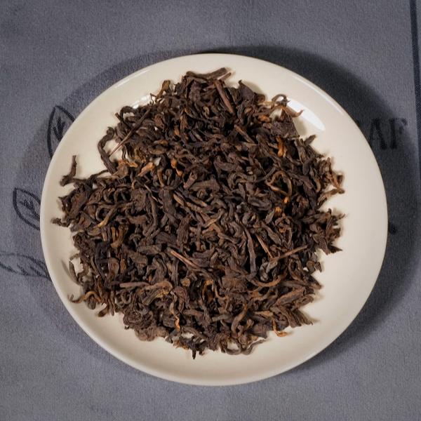 Close-up of 2014 Aged Liubao Dark Tea dry leaves on a white saucer, showcasing its dark, rich appearance with hints of golden hues, indicative of its Betel Nut Aroma and Cooling Finish.