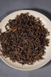 Close-up of 2014 Aged Liubao Dark Tea dry leaves on a white saucer, showcasing its dark, rich appearance with hints of golden hues, indicative of its Betel Nut Aroma and Cooling Finish.