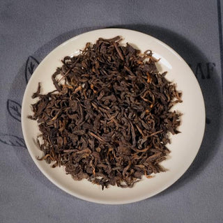 Close-up of 2014 Aged Liubao Dark Tea dry leaves on a white saucer, showcasing its dark, rich appearance with hints of golden hues, indicative of its Betel Nut Aroma and Cooling Finish.