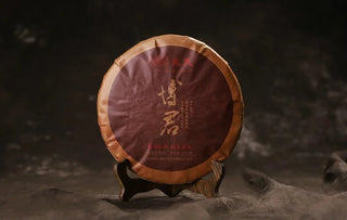 Front view of the 2017 Mengku Rongshi Bojun Ripe Pu-erh 500g cake, dark red paper wrap with gold calligraphy, on a wooden stand.
