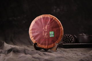 Front view of the 2019 Mengku Rongshi Bojun Ripe Pu-erh 500g cake, dark red paper with gold calligraphy, on a wooden stand.