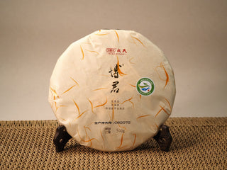 Front of the 2020 Mengku Rongshi Bojun Ripe Pu-erh 500g cake, rice paper with orange silk decoration, on a wooden stand.