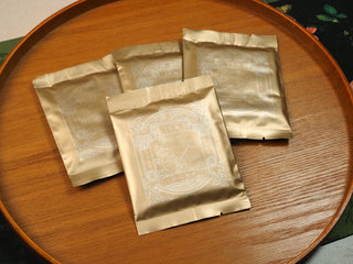 Four sealed sachets of 2024 sun-dried Fuding Shou Mei and Chen Pi waffle cakes stacked on a wooden round tray, gold tea label on packaging.
