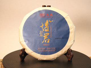 Front view of the 500g Mengku Rongshi Bojun Ripe Pu-erh 7.0 (2026) cake, blue paper wrap with gold calligraphy, on a wooden stand.