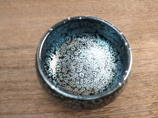 Top view of a Da Chan Ding Jianzhan tea bowl with dense silver oil-spot crystals and blue-green iridescence on a black glaze, resting on a wooden surface.