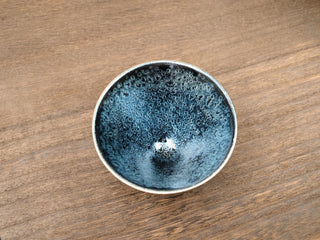 Top-down view of a Da Dou Li sky eye oil spot Jianzhan tea bowl on a wooden table, showing densely packed silver-blue crystals across a deep black interior.