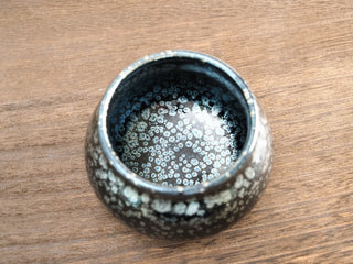 Top view of a Fo Du Jianzhan tea bowl with silver oil-spot crystals on a black glaze, resting on a wooden surface.