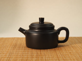 Heini Dezhong Yixing teapot in raw black clay, front side view with upright spout and flat stud knob on a bamboo mat