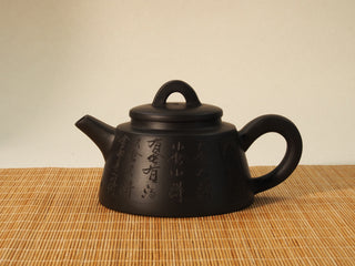 Black Zhu Ni Shede Yixing teapot in side view, engraved calligraphy, arched knob lid, on bamboo mat background