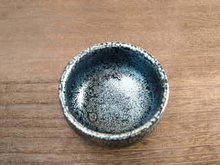 Top view of a Jiang Jun Bei Jianzhan tea bowl with silver oil-spot crystals on a black glaze, resting on a wooden surface.