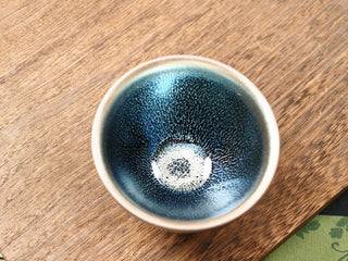Top view of a Lan Qi Lin Shu Kou Jianzhan tea bowl on a wooden table, showing a small pale central eye with fine silver pinpricks radiating outward on a black-blue ground.