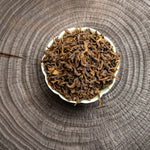 Dry leaves of Mang Fei 2022 loose leaf ripe pu-erh tea displayed in a white ceramic dish on a dark wooden tea tray.