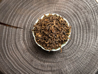 Dry leaves of Mang Fei 2022 loose leaf ripe pu-erh tea displayed in a white ceramic dish on a dark wooden tea tray.