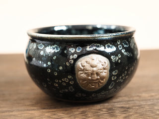 Front view of a Rui Shou sky eye oil spot Jianzhan tea bowl on a wooden table, showing the rounded belly and a raised unglazed clay auspicious beast medallion on the body.