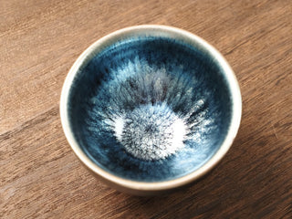 Top view of a Shu Kou frosted hundred-flower Jianzhan tea bowl on a wooden table, showing a soft blue-silver crystalline bloom across a dark blue-black interior with a brighter silver rim.