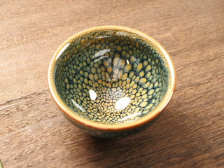 Top view of a Shu Kou green partridge Jianzhan tea bowl on a wooden table, showing dense olive-green partridge-feather speckles across the dark interior and a warm orange-gold rim band.