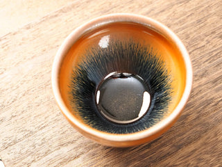 Top view of a Shu Kou hare's-fur Jianzhan tea bowl on a wooden table, showing dense dark brown-to-black streaks radiating from the center of a deep dark interior, with a warm amber rim.