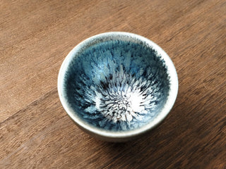 Top view of a Shu Kou hundred-flower Jianzhan tea bowl on a wooden table, showing silver-blue crystalline speckles radiating from the center across a dark blue-black interior, with a bright silver-blue rim.