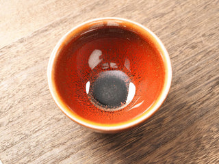 Top view of a Shu Kou red hare's-fur Jianzhan tea bowl on a wooden table, showing fine dark brown-to-black streaks radiating from a deep near-black center across a red-orange interior, with a bright orange-gold rim.