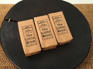 Three kraft-wrapped The Little Brick fu brick tea packs with OrientaLeaf x Genshe co-branded labels on a black wooden tray