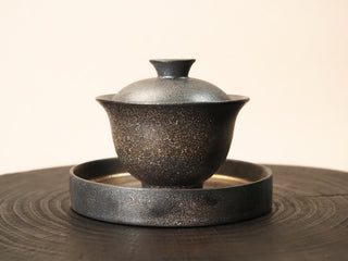 Wood-fired stoneware gaiwan with tea tray from Dehua Fujian, side view on a wooden cross-section, rough textured kiln-change glaze.