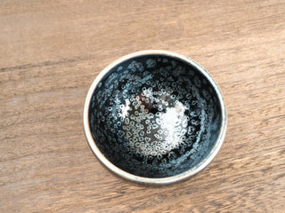 Top view of a Wu Zu Bo sky eye oil spot Jianzhan tea bowl on a wooden table, showing a large central pale crystal cluster surrounded by smaller silver rings on a deep black interior.