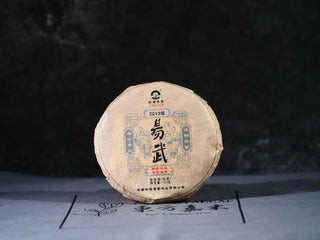 Front view of Yiwu Raw Pu-erh Cake – 2013 Spring Tea, Hand-Pressed 2023, Sweet & Layered (100g) with packaging, showing the round cake shape and elegant tea wrapper
