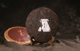 Unwrapped 2017 Bojun 500g cake, dark fermented leaves with golden buds, the inner ticket label visible, on a wooden stand.