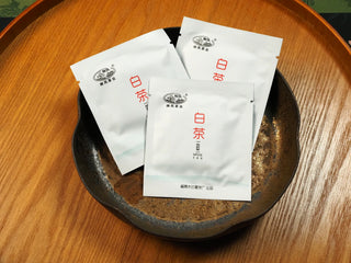 Three sealed sachets of 2018 sun-dried Fuding White Peony compressed tea biscuits, each marked with white tea label, displayed on a dark ceramic plate.