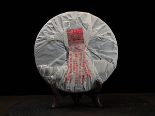 Back view of the 2021 Dayi 7572 ripe puerh tea cake wrapped in traditional paper, displayed on a wooden stand.
