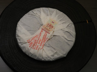 Traditional paper-wrapped back view of the 2019 Dayi 7572 ripe puerh tea cake, displayed on a wooden stand.