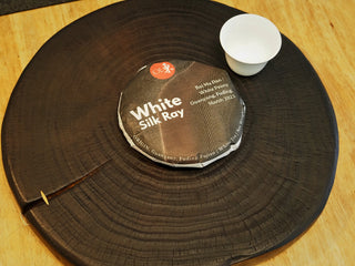 Wrapped Silk Ray white tea cake showcased on a dark charcoaled wooden presentation board next to a white porcelain teacup.