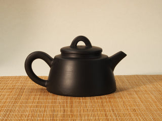 Black Zhu Ni Shede Yixing teapot reverse side view with smooth plain body, handle and spout, sitting on a bamboo mat background