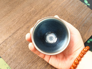 Handheld top-down view of a Lan Qi Lin Shu Kou Jianzhan bowl in palm, showing the full interior with fine silver-blue pinpricks and a small pale central cluster.