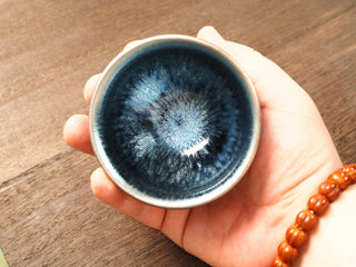 Handheld top-down view of a Shu Kou frosted hundred-flower Jianzhan bowl in palm, showing the soft matte blue-silver crystalline bloom radiating from a deep blue-black center toward a silver-blue rim.