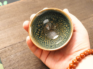 Handheld top-down view of a Shu Kou green partridge Jianzhan bowl in palm, showing the green ground covered in small, near-touching yellow-green to amber partridge-feather speckles.