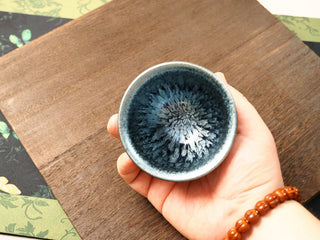 Handheld top-down view of a Shu Kou hundred-flower Jianzhan bowl in palm, showing the silver-blue crystalline burst radiating from a deep blue-black center toward a brighter blue rim.