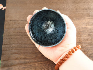 Handheld top-down view of a Shu Kou sky eye oil spot Jianzhan bowl in palm, with blue-green iridescent crystals around a central cluster.