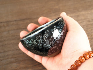 Handheld side view of a Wu Zu Bo sky eye oil spot Jianzhan bowl, showing the deep open form and silver-ring oil-spot crystals on the exterior.