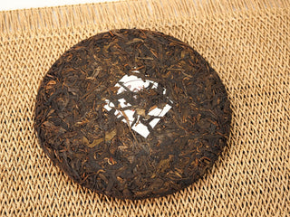 Front of unwrapped Jing Chang Hao Quiet Years 2018 raw Pu-erh 357g cake, dark Bulang arbor leaves with white nei fei label on a woven mat