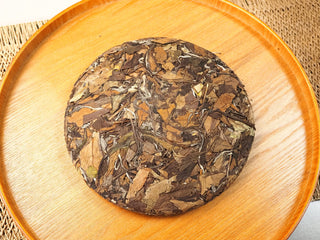 Top view of 2018 sun-dried Fuding Shou Mei 350g compressed cake on a round wooden tray, dark leaves with silvery stems pressed firmly.