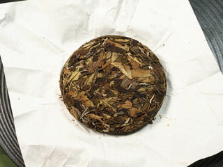 Fortune 2021 sun-dried Fuding Shou Mei 50g mini cake unwrapped, top view of loosely pressed dark leaves with silvery buds.