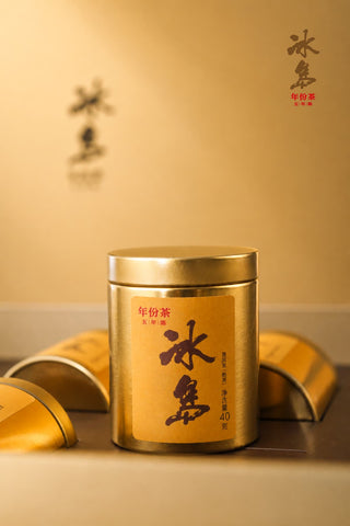 Single golden tin of Mengku Rongshi Bingdao Aged Ripe Pu-erh coin cakes (40g) on a dark surface, with other tins blurred behind.