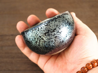 Handheld side view of a Bu Dao Weng sky eye oil spot Jianzhan bowl, showing the rounded belly covered in raised silver oil-spot crystals.