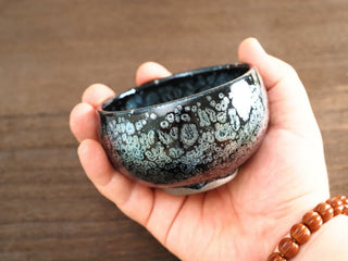 Side view of a Da Chan Ding Jianzhan tea bowl held in hand, showing the low broad form and large raised silver oil-spot crystals on the dark exterior.