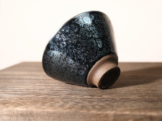 Side view of a Da Dou Li sky eye oil spot Jianzhan bowl tilted up on its foot, showing the conical silhouette and dense silver oil-spot pattern.
