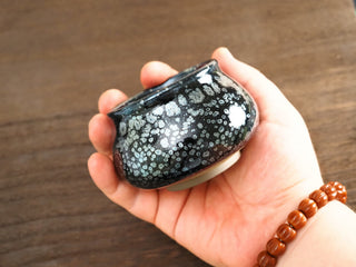 Side view of a Fo Du Jianzhan tea bowl held in hand, showing the rounded Buddha's belly form and silver oil-spot pattern on the dark exterior.