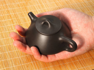 Hand holding a Black Zhu Ni Ziye Shipiao Yixing teapot in the palm, showing its small 130ml size and bridge knob lid