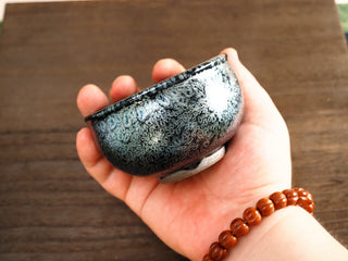 Side view of a Jiang Jun Bei Jianzhan tea bowl held in hand, showing silver oil-spot pattern and faint blue-green iridescence on the dark exterior.