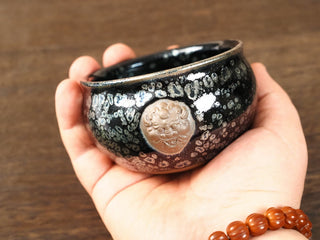 Handheld side view of a Rui Shou sky eye oil spot Jianzhan bowl, showing a raised unglazed clay auspicious beast medallion against the dark glazed body.